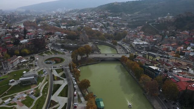 Aerial drone flyover of Tbilisi, Georgia featuring Kura River, futuristic Peace Bridge area, Rike Park, and modern architecture contrasting with historic cityscape.