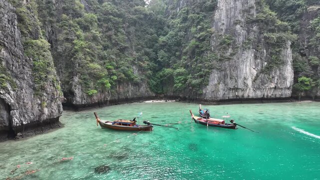 Aerial view of longtail boats in crystal-clear turquoise waters surrounded by limestone cliffs covered in lush green vegetation, Kho Phi Phi Island, Thailand.