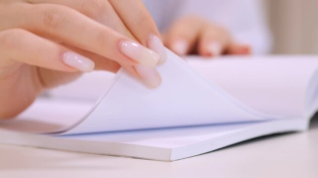 Hand with polished nails flips through blank notebook pages. Smooth white paper turns under careful fingers. Soft focus highlights gentle motion. Clean desk surface visible beneath book