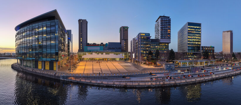 A wide aerial panorama of the MediaCityUK waterfront plaza in Salford Quays, showcasing the headquarters of BBC North and the ITV studios during the blue hour.