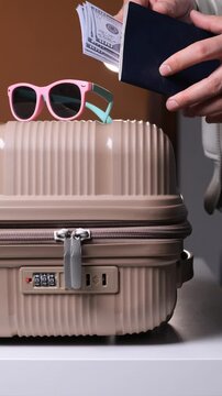 Vertical video traveler arranging cash inside passport beside suitcase and sunglasses, preparing finances and essentials for international trip