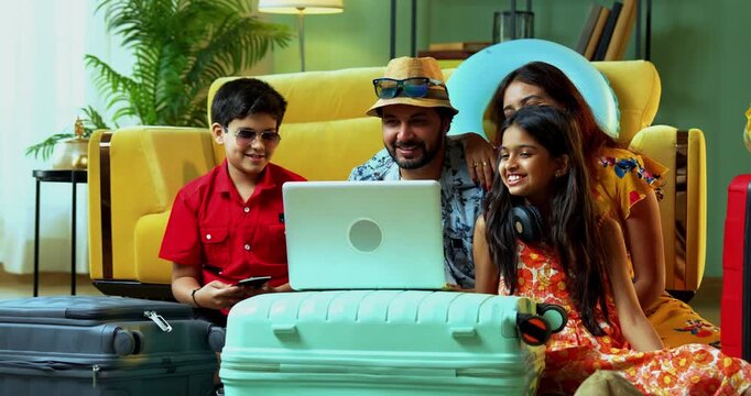 Cheerful Indian family of four sitting together at home. Using a laptop to book a hotel for their summer vacation trip with suitcases and passports ready
