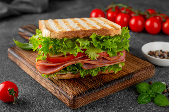 Ham and cheese sandwich with fresh lettuce and tomato on toasted bread. Close-up on a wooden board, juicy layers, deli style. Healthy fast food, dark background, selective focus.
