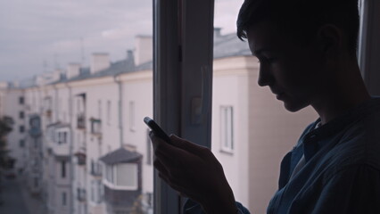 Fototapeta premium Young Man Using Smartphone Indoors by Window at Dusk, have fun and enjoy