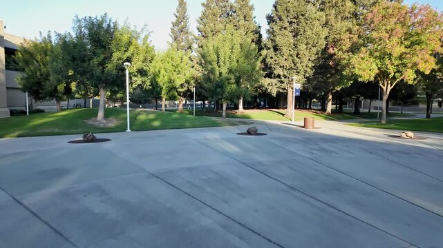 First-person perspective walking through the grounds of Modesto Junior College in California, showing a paved courtyard, green lawns, and lush trees on a sunny morning