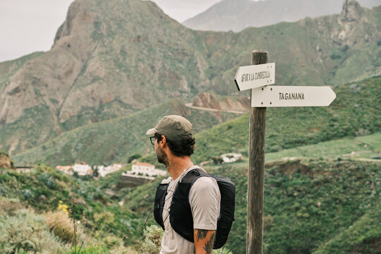 A male hiker in a cap and backpack stands at a signpost, gazing at the lush green mountainous landscape and distant village. Ready for adventure and exploration.