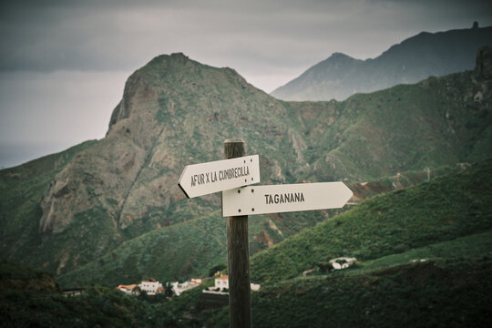 Rustic signpost directs towards "AFUR X LA CUMBRECILLA" and "TAGANANA" in a stunning mountain range. Lush green slopes and a dramatic cloudy sky create a picturesque backdrop, hinting at remote villag