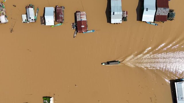 Aerial view of houses with tin roofs stand on stilts in the muddy water, as a boat cuts through the water, Chong Kneas, Cambodia.
