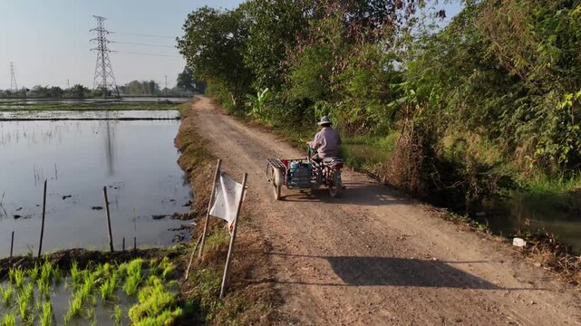 Aerial view of a person riding a three-wheeled vehicle on a dirt path, contrasting with the still water and lush greenery along the roadside, Chiang Rai, Thailand.