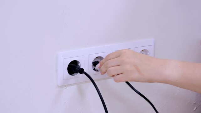 Hand plugs white cord into wall socket. Three black cords already plugged in. Simple, clean wall socket setup. Minimalist, bright background. Realistic, everyday home scene