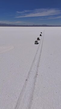 Aerial view of vehicles leaving tracks on the vast, bright white salt flats contrasting with the blue sky and distant mountains, Salar de Uyuni, Bolivia.