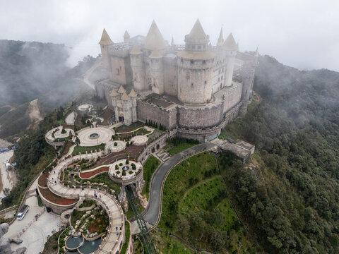 Aerial view of imposing castle piercing through a veil of mist, a medieval fortress amidst lush greenery, Ba Na Hills, Vietnam.