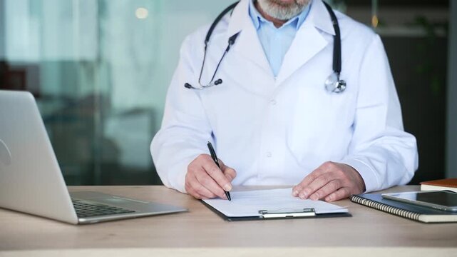 Close-up of male doctor hands signing medical documents on desk. Mature physician in white coat writing prescription and filling out patient report on clipboard, reviewing report at hospital office.