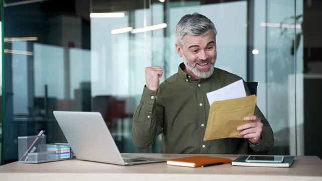 Happy gray-haired businessman sitting at office desk reading letter with great news. Excited mature man smiling with joy, feeling successful and satisfied with positive business results at workplace.