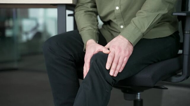 Close-up of male hands holding and massaging knee while sitting at office desk. Businessman suffering from severe knee pain, rubbing leg muscles, feeling discomfort, cramps or rheumatism at workplace