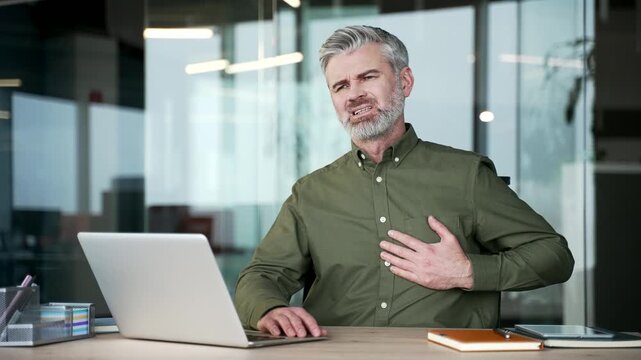 Gray-haired businessman sitting at office desk suffering from heart attack. Sick mature man feeling sudden chest pain, holding his chest with hands, experiencing tension or heart problem at workplace.