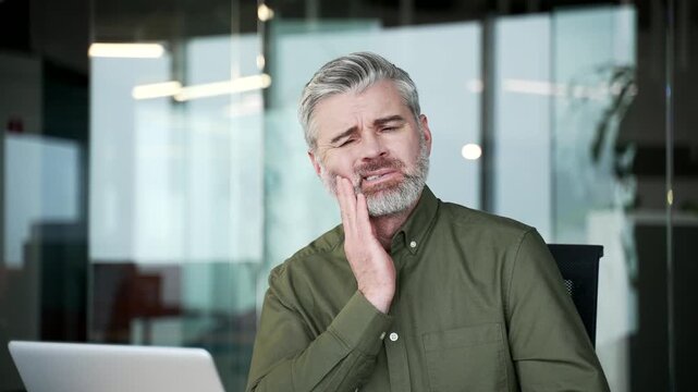 Close-up of gray-haired businessman suffering from toothache. Sick mature man holding hand on cheek, feeling pain and discomfort caused by dental problem while sitting at modern office workplace.
