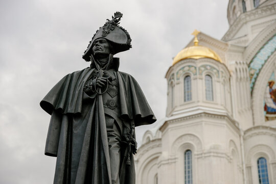 Bronze statue of Russian Admiral Fyodor Ushakov in a tricorne hat and cloak, standing before the St. Nicholas Naval Cathedral with a gold dome in Kronstadt on a cloudy day.