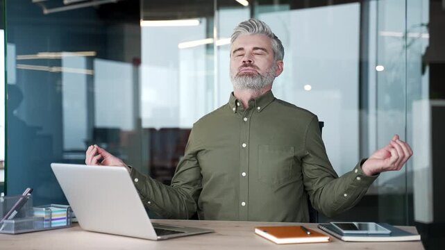 Gray-haired businessman sitting at office desk meditating with closed eyes during break at work. Mature man relaxing, relieving stress and finding balance while practicing mindfulness at workplace.