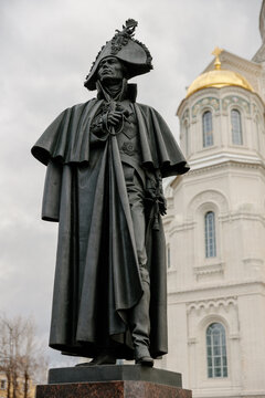 Bronze statue of Russian Admiral Fyodor Ushakov in a tricorne hat and cloak, standing before the St. Nicholas Naval Cathedral with a gold dome in Kronstadt on a cloudy day.