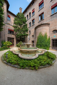View of a tranquil fountain surrounded by lush greenery in a courtyard framed by aged brick buildings with red accents, casting gentle shadows, Ravenna, Italia.