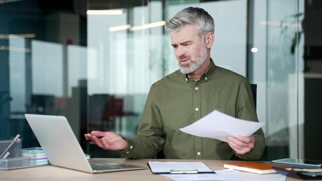 Confused mature businessman sitting at desk in office, holding document and staring at laptop screen with puzzled expression, struggling to understand financial report and data, uncertainty and stress
