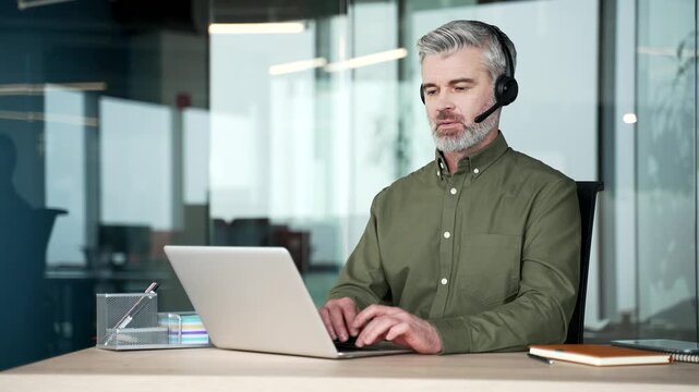 Portrait of mature gray-haired businessman wearing headset and working on laptop in modern office then looking confidently at camera, professional operator customer service and corporate communication