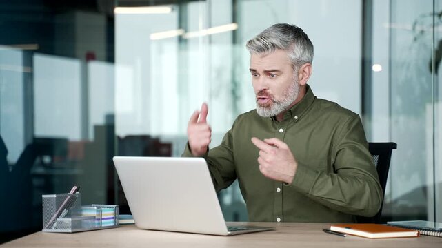 Angry mature businessman sitting at desk in office, arguing with subordinate during online meeting on laptop, expressing frustration and dissatisfaction, shouting on video call, gesturing emotionally