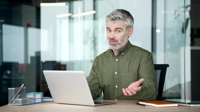 Mature gray-haired businessman struggling with laptop in modern office, looking at screen in confusion and then glancing at camera, feeling frustrated and puzzled after technical problem during work