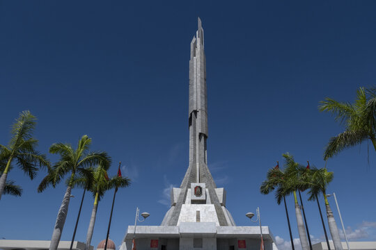 View of the towering, modern Agostinho Neto Memorial piercing the vibrant blue sky, framed by swaying palm trees in the warm Angolan sun, Luanda, Angola.