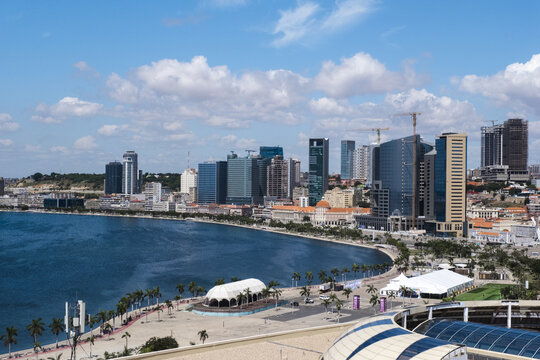 View of the modern city skyline reflected in the bay's waters, with palm trees lining the shore under a bright sky, in Luanda, Angola.