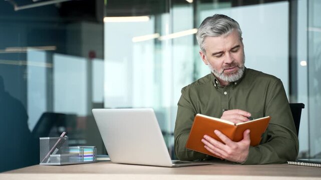 Gray-haired businessman watching video call conference on laptop and taking notes in notebook at workplace. Manager attending remote online training, focused on learning, professional tasks in office.