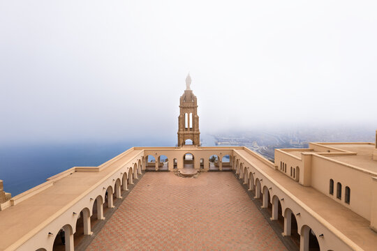 View of a tan building with arched walkways and a tower shrouded in mist, overlooking the serene blue sea, creating a tranquil scene, Oran, Algeria.