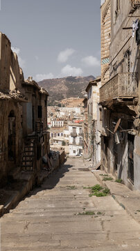 View of sun-drenched stone steps descending through aged buildings towards distant hills under a hazy sky, creating a captivating urban scene, Oran, Algeria.