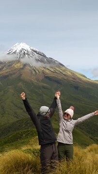 Couple celebrates reaching a mountain viewpoint with Mount Taranaki behind them, vertical static shot.