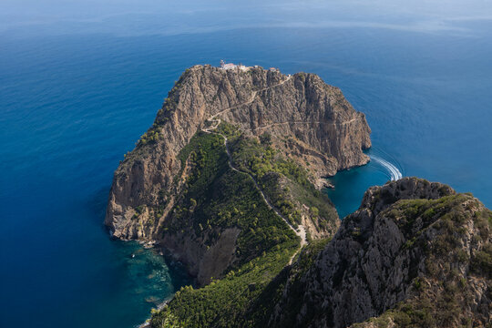 View of rugged cliffs meet the vast, azure sea, green vegetation contrasting with rocky terrain under a clear sky, a boat races by, Bejaja, Algeria.
