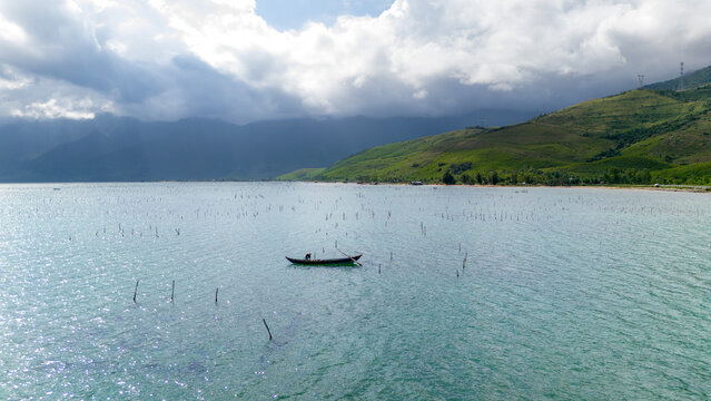 Aerial view of a lone boat amidst a serene lagoon, the oyster farm contrasting with the distant green hills under a cloudy sky, tt. Lang Co, Hue, Vietnam.