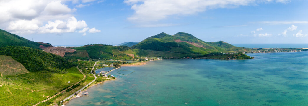 Aerial view of verdant hills meet the turquoise lagoon, fringed by a coastal road and oyster farms under a vast sky, tt. Lang Co, Hue, Vietnam.
