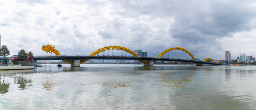 View of the golden Cau Rong bridge, a serpentine dragon of steel and light, arches over the tranquil river under a sky thick with clouds, Da Nang, Vietnam.