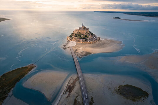 Aerial view of Mont Saint-Michel, a medieval abbey perched on a tidal island, connected by a causeway amidst the swirling blues and sandy textures of the surrounding waters, Mont Saint Michel, France.