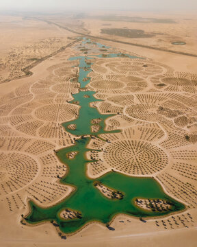 Aerial view of a vibrant green oasis cuts through the stark desert landscape, with circular patterns etched into the sand, a striking contrast of nature and design, Dubai, United Arab Emirates.