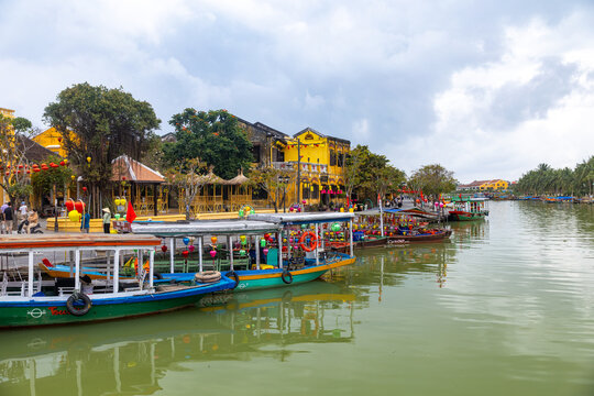 Hoi An, Vietnam - 28 January 2026: View of boats lined along the riverbank in Hoi An's Old Town, their vibrant colors reflecting in the calm water under a cloudy sky.