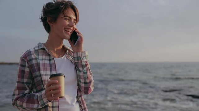 Young happy woman wears shirt casual clothes talk speak on mobile cell phone drink coffee rest relax in free time walk on sea ocean sand shore beach outdoor seaside in summer day. Lifestyle concept