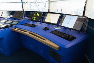 A modern ship bridge featuring a blue navigation console with multiple monitors showing radar and electronic charts, keyboards and control panels used for maritime navigation and vessel monitoring © evannovostro