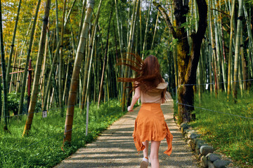 Fototapeta premium Woman running along a path in a bamboo forest, back view wearing an orange skirt, summer nature scene with green trees, motion blur and sunlight on a peaceful outdoor walkway.