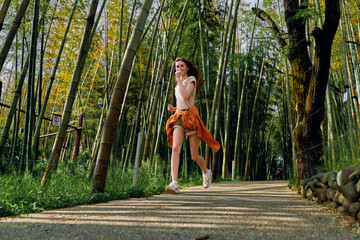 Fototapeta premium Woman running along a bamboo forest path, smiling and jogging with smartphone and headphones. Casual outfit with shorts and sweater tied around waist during outdoor fitness session.