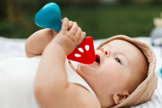 Beautiful happy baby in hat lying on blanket and playing with teether on background of sunny summer garden. Childhood. Portrait of cute infant relaxing in park, enjoying outdoors