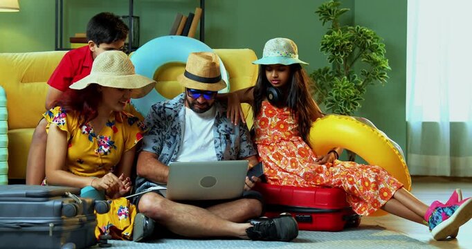 Happy Indian family of four with laptop and suitcases in modern home doing web check-in or booking an international holiday trip together in summer attire