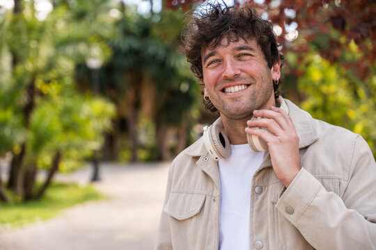 Smiling young man wearing headphones around his neck, looking at the camera in a park with lush greenery