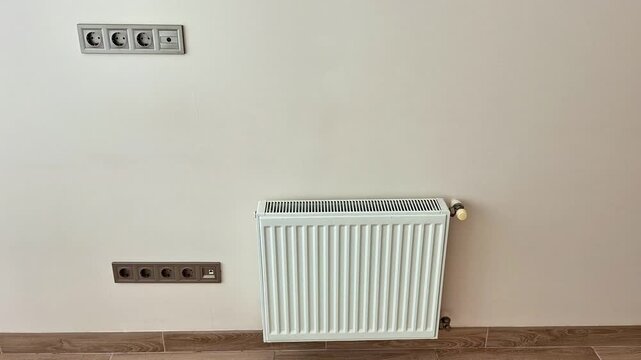 White wall mounted radiator with valve positioned above tiled floor, paired with two sets of electrical outlets, showing heating equipment and utility placement in a room